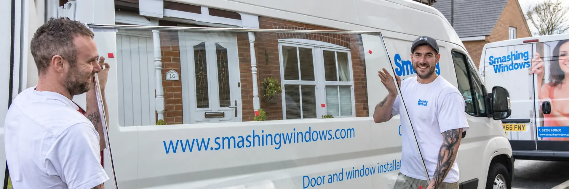 Two Smashing Windows fitters carrying a replacement sealed glass unit beside the company's branded vans